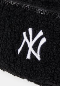 Svart fluffig tyg med en framträdande vit broderad "NY"-logotyp. Har en svart dragkedja längs den övre kanten. Mjuk textur och rundad form.