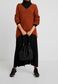 Rostfärgad stickad tröja med V-ringning och långa ärmar, kombinerad med en svart lång kjol. Håller en svart bucket bag och bär svarta sneakers.