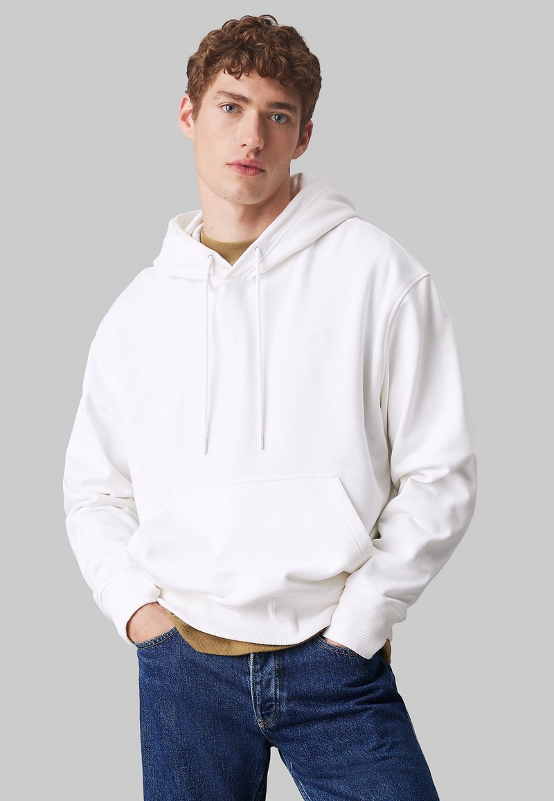 Junger Mann mit lockigem Haar, der einen weißen Kapuzenpullover über einem braunen Shirt und blauen Jeans trägt, mit teilweise in die Taschen gesteckten Händen, neutraler Hintergrund.