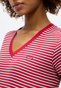 Femme portant un t-shirt rayé rouge et blanc avec un petit logo rouge Levi's sur la poitrine, cheveux bouclés visibles.
