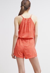 Czerwony kombinezon w wzory z cienkimi ramiączkami, z małymi białymi kształtami diamentów, zmarszczonym tyłem i luźnym krojem. Krótki fason szortów.