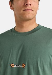 Green cotton t-shirt with short sleeves, round neck, and orange floral logo. Soft texture, relaxed fit, and minimalistic design.