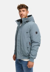Ung man med kort, lockigt hår iförd en ljusblå huvjacka för vintern och blå jeans, stående mot en enfärgad bakgrund.