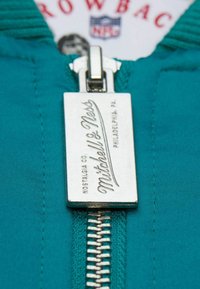 Teal Reißverschlusszug mit einem silbernen Metallschild, das mit "Mitchell & Ness" graviert ist. Verfügt über eine strukturierte Oberfläche und eine polierte Optik.