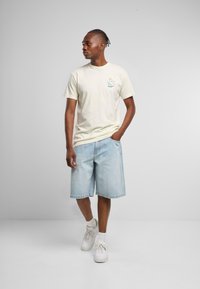 Lys gul bomulls-T-skjorte med et lite blått grafikkmotiv på brystet, sammen med løse, lyse denimshorts og hvite joggesko.