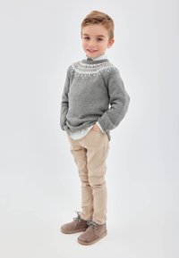 Suéter gris de punto con un canesú estampado, chinos beige y botines grises. El niño posa con las manos en los bolsillos frente a un fondo claro.