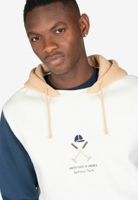 Sudadera con capucha de cuerpo crema, mangas azul marino y capucha beige. Presenta un gráfico de dos mazos cruzados y una gorra, con "Estados Unidos de América".