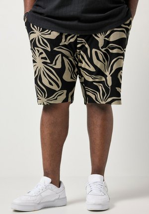 Ben iført sorte shorts med store beige bladmønstre, hvide sneakers og en sort skjorte med hænderne i lommerne.