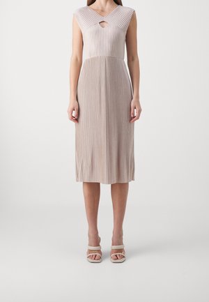 Kobieta w beżowej, plisowanej sukience midi bez rękawów z dekoltem w serek i małym wycięciem z przodu, w połączeniu z beżowymi sandałami na obcasie odsłaniającymi palce.