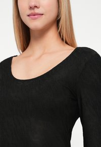 Haut à manches longues noir avec un motif texturé, doté d'un décolleté large en forme de scoop et en tissu doux, présenté sur un fond neutre.