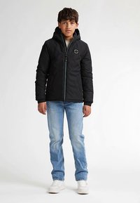 Svart vadderad jacka med dragkedja, huva och logodetalj, bärs över en grå tröja, kombinerad med blå jeans och vita sneakers.