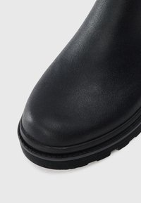 Bout en cuir noir avec surface texturée et semelle en caoutchouc épaisse sur un fond blanc.
