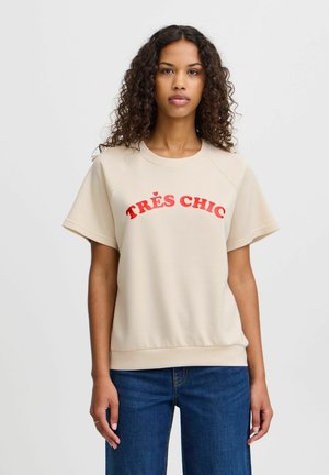 Sweat-shirt beige à manches courtes avec un col rond côtelé et "TRÈS CHIC" en lettres rouges en gras sur le devant. Tissu lisse, coupe décontractée.