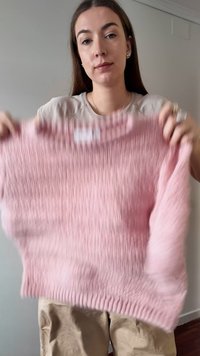 Femme aux longs cheveux bruns tient devant elle un pull en tricot rose doux, sur un fond de mur uni.