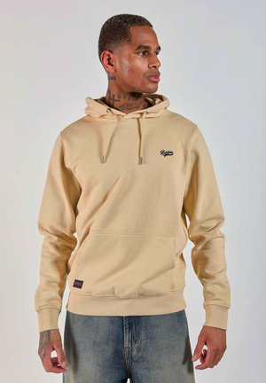 Homme avec des tatouages sur le cou et les mains portant un hoodie beige et un jean bleu, regardant vers la droite sur un fond uni.