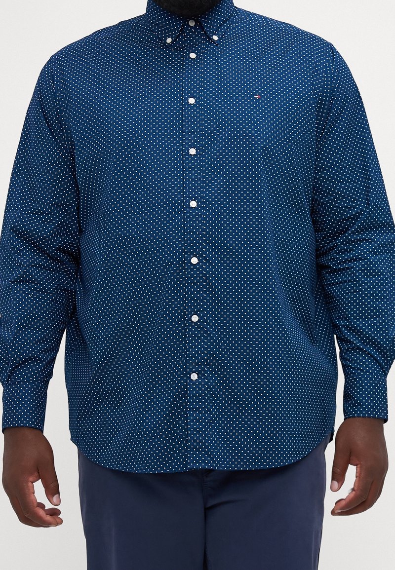 Homme portant une chemise bleu marine à manches longues boutonnée avec de petits pois blancs et un pantalon bleu marine assorti.
