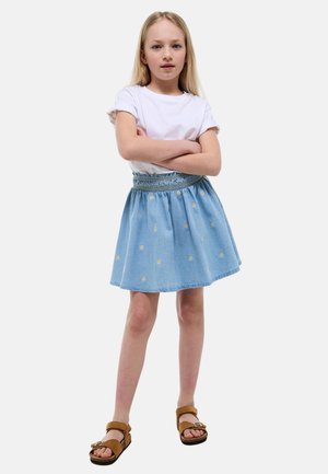 Jeune fille aux bras croisés portant un t-shirt blanc, une jupe bleue à imprimé marguerites et des sandales marron, debout devant un fond uni.