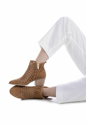 Gambe in pantaloni bianchi che indossano stivaletti beige intrecciati con tacco largo, un piede appoggiato sull'altro su uno sfondo bianco.