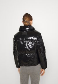 Sort pufferjakke med en blank finish, høj krave og et logo patch på bagsiden. Har brede quiltede sektioner og elastiske manchetter.