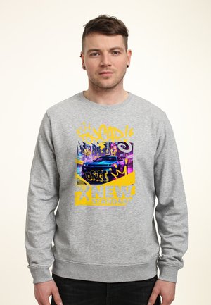 Mężczyzna noszący szarą bluzę z kolorową grafiką niebieskiego samochodu sportowego, żółtym tekstem graffiti i napisem "NEW STANDARD" z przodu.