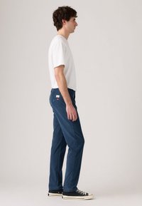 Tânăr stând lateral, îmbrăcat cu un tricou alb, pantaloni drepti albaștri și adidași negri cu talpă joasă, pe un fundal simplu.