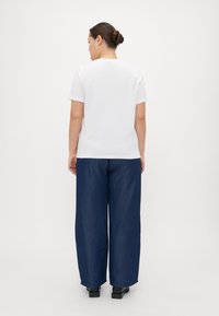 Hvit kortermet T-skjorte sammen med løst sittende, mørkeblå denimbukser; modell vender bort, og viser en enkel design og avslappet passform.