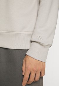 Světle šedá mikina s žebrovanými manžetami a lemem; obsahuje malou vyšitou nášivku ve stejném odstínu na rukávu. Jemná textura látky.