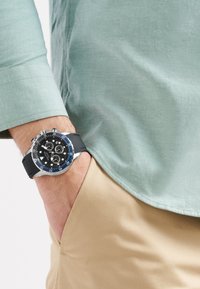 Melna hronogrāfa pulksteņa ar sudraba akcentiem, zilu rāmīti un gumijas siksniņu, kas valkāta uz rokas, kas iegrābta bēšās biksēs. Redzama gaiši zaļa krekla daļa.