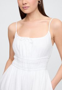 Vit klänning av lätt tyg med tunna axelband, knytning fram och insvängd midja. Slät textur med en flödande kjol.