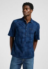 Camisa de manga corta de color azul oscuro con un patrón de cuadros, que presenta un cierre de botones en la parte delantera, un bolsillo y un cuello, fabricada con una tela texturizada.