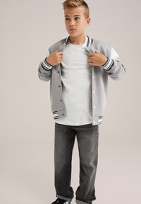 Chico ajustando una chaqueta de varsity gris y blanca, vistiendo una camiseta blanca, jeans oscuros y zapatillas negras, frente a un fondo claro y liso.