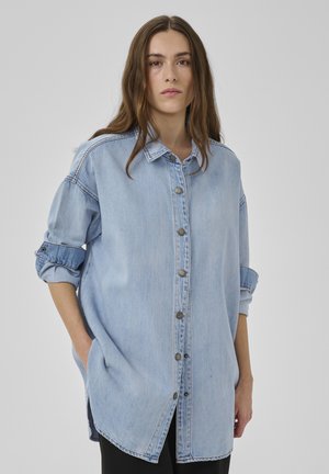 Mladá žena v dlhej svetlomodrej rifľovej košeli s vyhrnutými rukávmi stojí pred jednoduchým sivým pozadím.