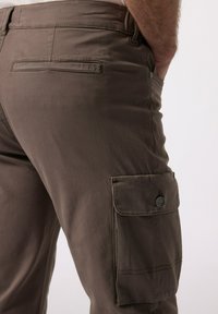 Hnědé cargo kalhoty vyrobené z lehkého materiálu, s boční kapsou se klopou a knoflíkem, a hladkým, mírně texturovaným povrchem.