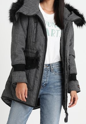 Grau kariertes Parka-Jackett mit einem mit Fell besetzten Kapuze, Reißverschluss, schwarzen Akzenten und Seitentaschen. Kombiniert mit einem weißen Hemd und blauen Jeans.
