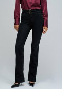 Jeans negros de pierna recta con una cintura alta, que cuentan con un cinturón de doble hebilla metálica y combinados con una blusa de satén color burdeos oscuro.