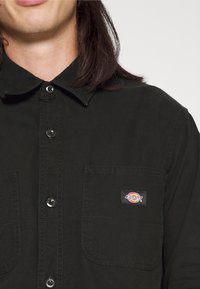 Camisa negra de botones con dos bolsillos frontales, un cuello clásico y un pequeño parche con el logo, hecha de un tejido duradero con un acabado mate.