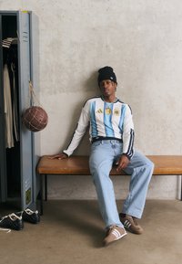 Ung man i blå jeans och randig sporttröja sitter på en bänk bredvid ett öppet skåp med sneakers och en hängande basketboll.