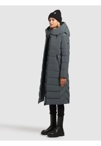 Manteau d'hiver long et gris en duvet avec un col haut, un motif de couture diagonal, une fermeture éclair à l'avant et des poches latérales. Porté avec des bottines noires et un bonnet.