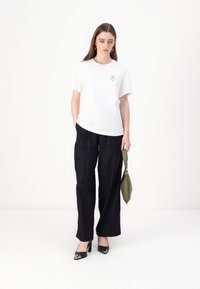 White cotton t-shirt with a turquoise graphic design, paired with black pinstripe wide-leg trousers and black heeled shoes. Holding a green bag.
