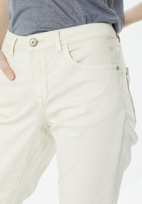 Pantalones de mezclilla de color beige claro con un diseño ajustado, que cuentan con un botón en la parte frontal, bolsillos laterales y detalles de costura sutiles. Textura suave en todo el tejido.