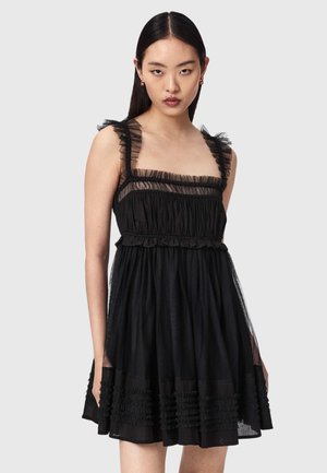 Kobieta z prostymi czarnymi włosami, ubrana w czarną, bezrękawną sukienkę z marszczonymi przezroczystymi ramiączkami i dołem, stojąca na jasnoszarym tle.