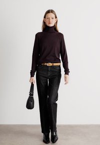 Pull à col roulé noir, jean slim noir, ceinture marron, sac à main noir et bottes noires à talons pointus. Le mannequin se tient devant un fond neutre.