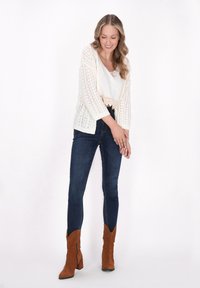 Cardigan all'uncinetto leggero di colore crema con maniche 3/4, abbinato a un top bianco corto, jeans blu aderenti e stivaletti alla caviglia in suede marrone.