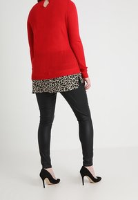 Pull rouge à manches longues avec une ouverture en forme de goutte dans le dos, superposé à un haut imprimé léopard noir et beige, associé à des leggings en cuir noir et des talons.