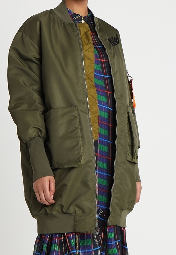 Blouson bomber vert olive avec fermeture éclair sur le devant, deux grandes poches, poignets côtelés et doublure matelassée, porté par-dessus une robe à carreaux.