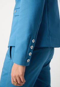 Veste de costume bleue au design structuré, avec une texture lisse, six boutons argentés sur les poignets et une coupe cintrée.