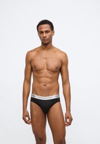 Μαύρα βαμβακερά σλιπ με γκρι λάστιχο που φέρει το λογότυπο "TOMMY HILFIGER", εφαρμοστή γραμμή και απαλό υλικό. Το μοντέλο ποζάρει όρθιο.