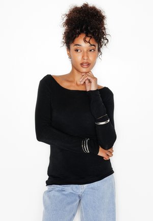 Donna con capelli ricci, indossa un top nero a maniche lunghe e jeans azzurri, posa con una mano sul mento e l'altra incrociata in vita.