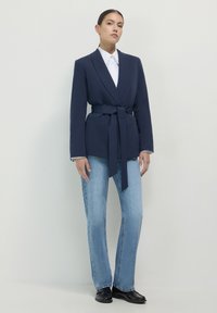 Donna che indossa un blazer blu scuro con cintura, camicia bianca, jeans azzurri e scarpe nere, in piedi contro uno sfondo semplice.
