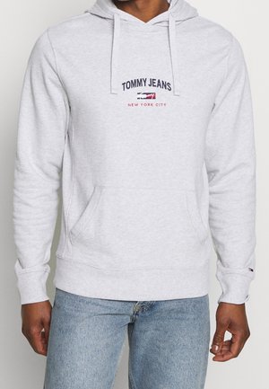 Jasnoszara bluza z kapturem z kieszenią z przodu i sznurkami, logo "Tommy Jeans New York City" na piersi, noszona z jasnoniebieskimi dżinsami.